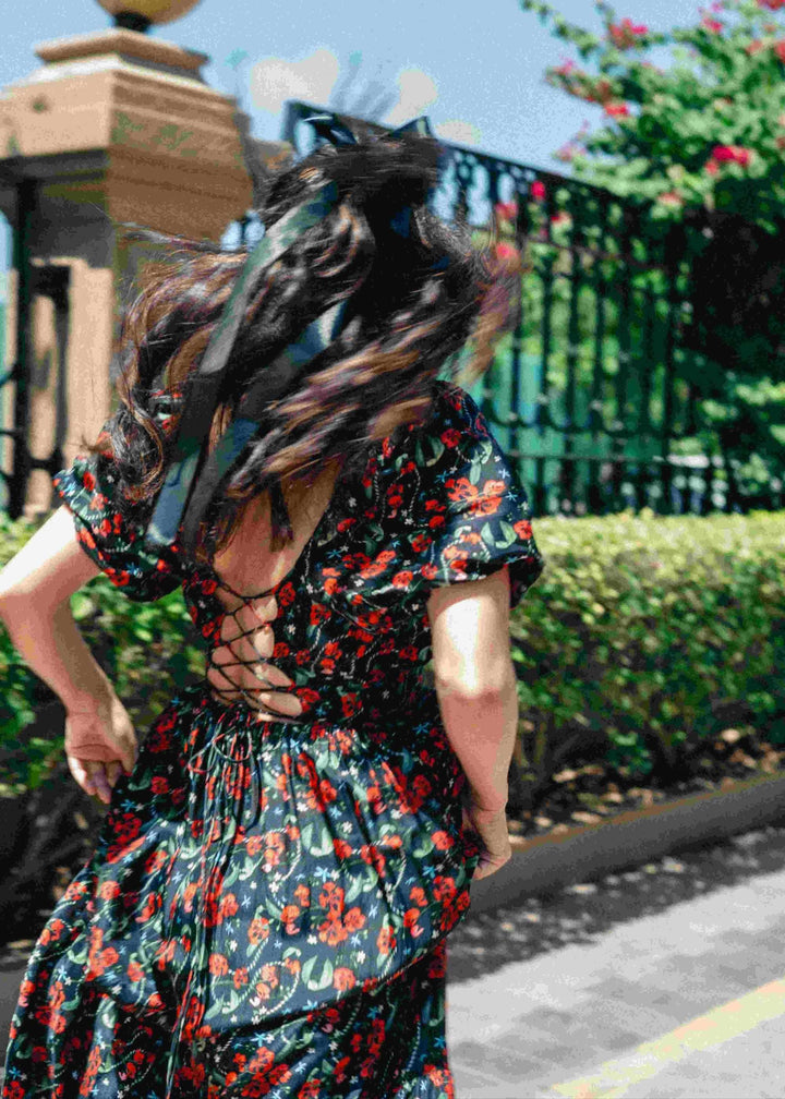A woman wearing a floral Royal Poinciana back tie-up corset dress with pockets, featuring a deep front neckline and elasticated sleeves, running through a gate with a flowy hemline. swim suit 
swimming suits for women
swimdress for women 
swimdresses for women 
bikini dress 
swim suits for ladies 
swimming dress for ladies 
monokinis 
women's swimdresses 
swimwear