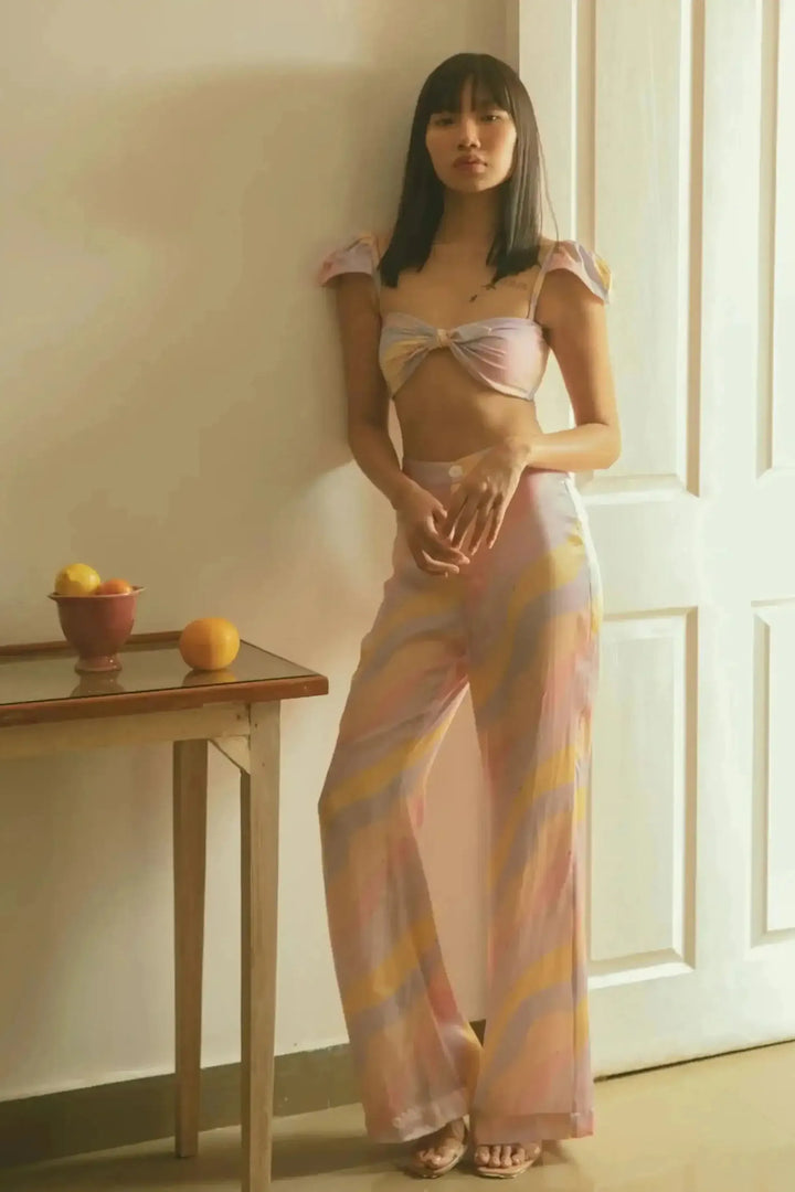 A model wearing a pink and orange striped bikini top with winged design and matching high waist trousers, standing in a room with a wooden table and chair in the background.